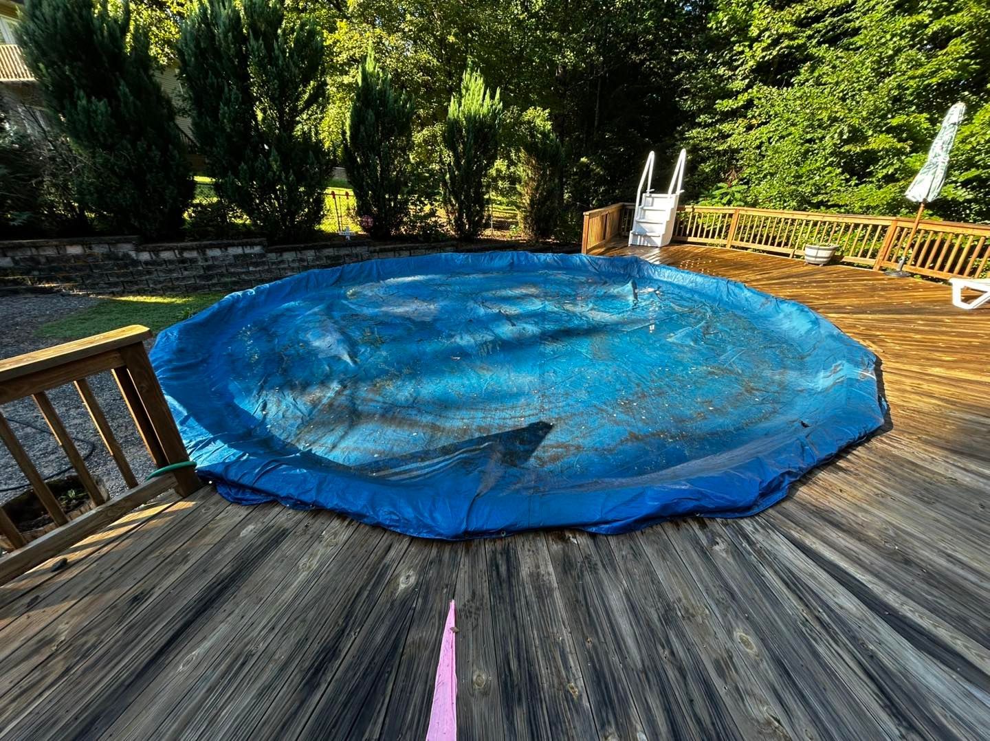 A round, above-ground swimming pool with a blue cover, set into a wooden deck in a backyard with trees in the background.