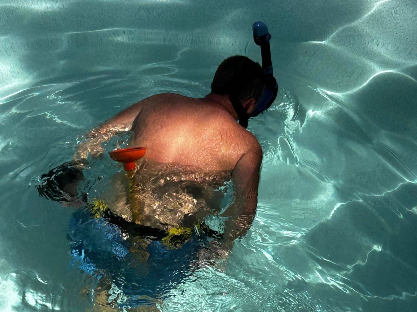 A person wearing a snorkel and mask swims in a pool with a bright orange plunger attached to their waist belt.