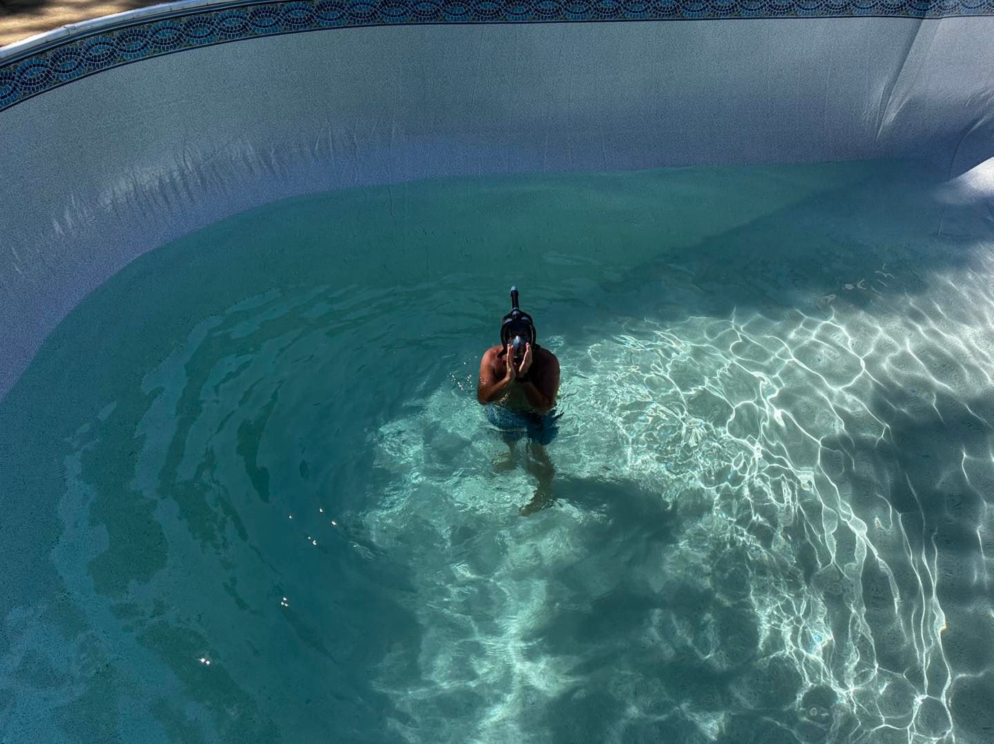 A person wearing a snorkel and mask stands waist-deep in the bright, clear blue water of a backyard swimming pool.