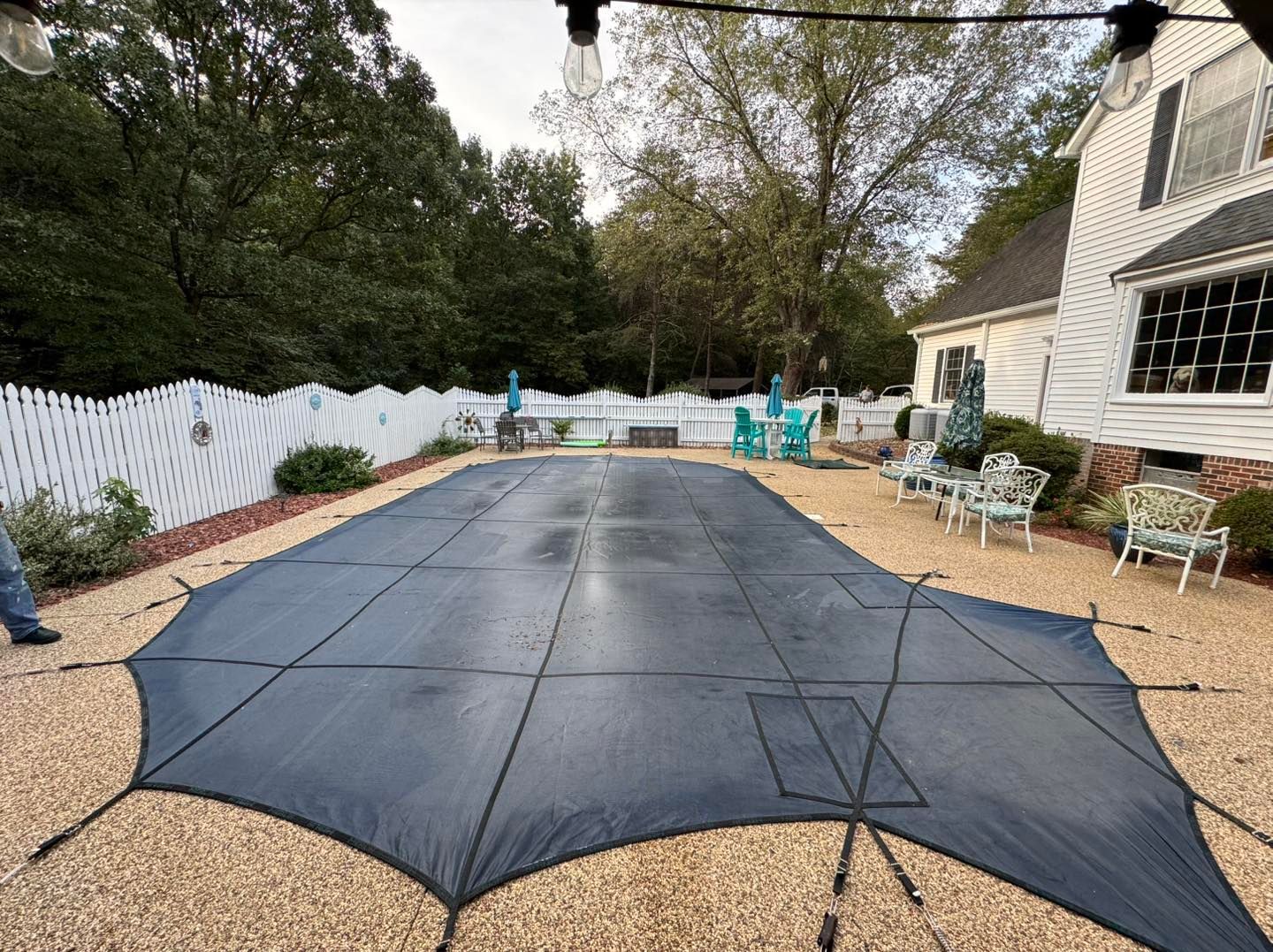 A dark, rectangular pool safety cover is anchored over a backyard swimming pool, surrounded by gravel and patio furniture.