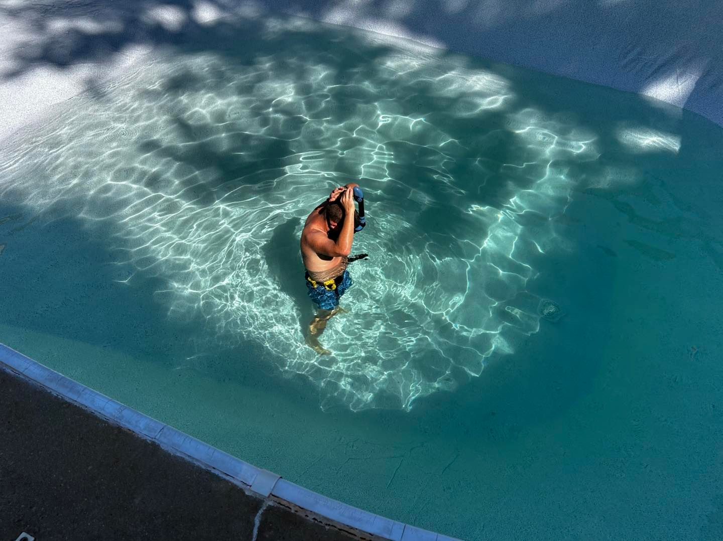 A person stands in a clear, bright blue swimming pool, adjusting a snorkel mask while water reflects sunlight.