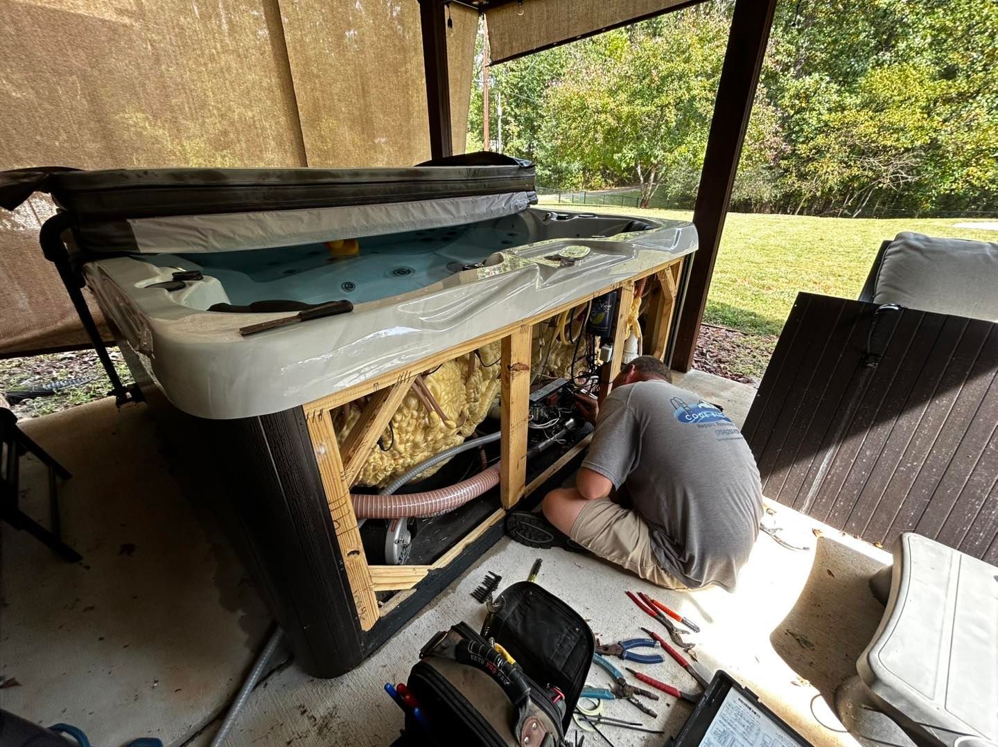 A person in a gray shirt repairs the exposed mechanical components of a hot tub on a concrete patio.