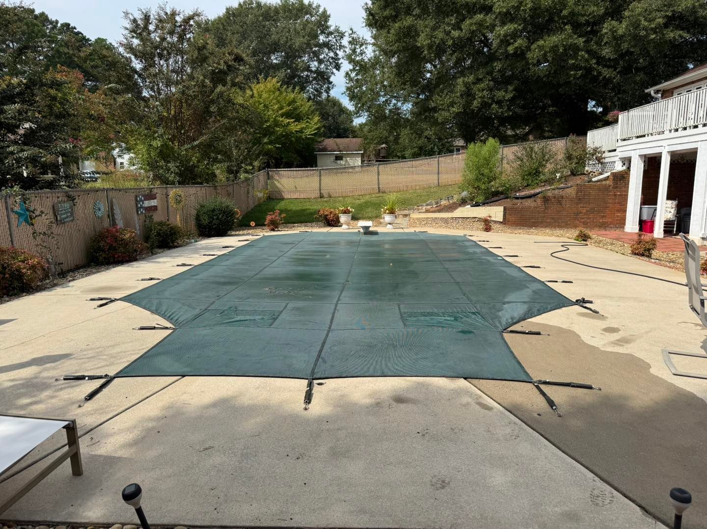 A rectangular swimming pool covered with a dark green safety mesh, set in a backyard patio surrounded by trees and a wall.