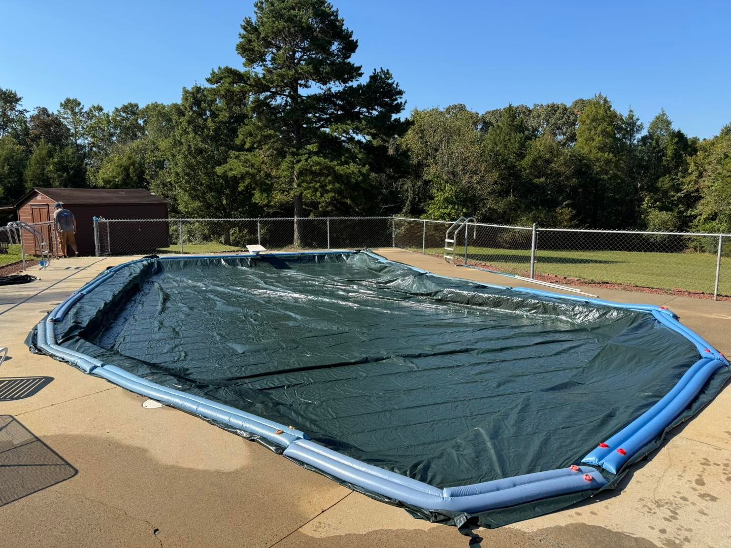 A rectangular swimming pool covered with a dark green tarp and secured by blue water-filled tubes on a concrete patio.