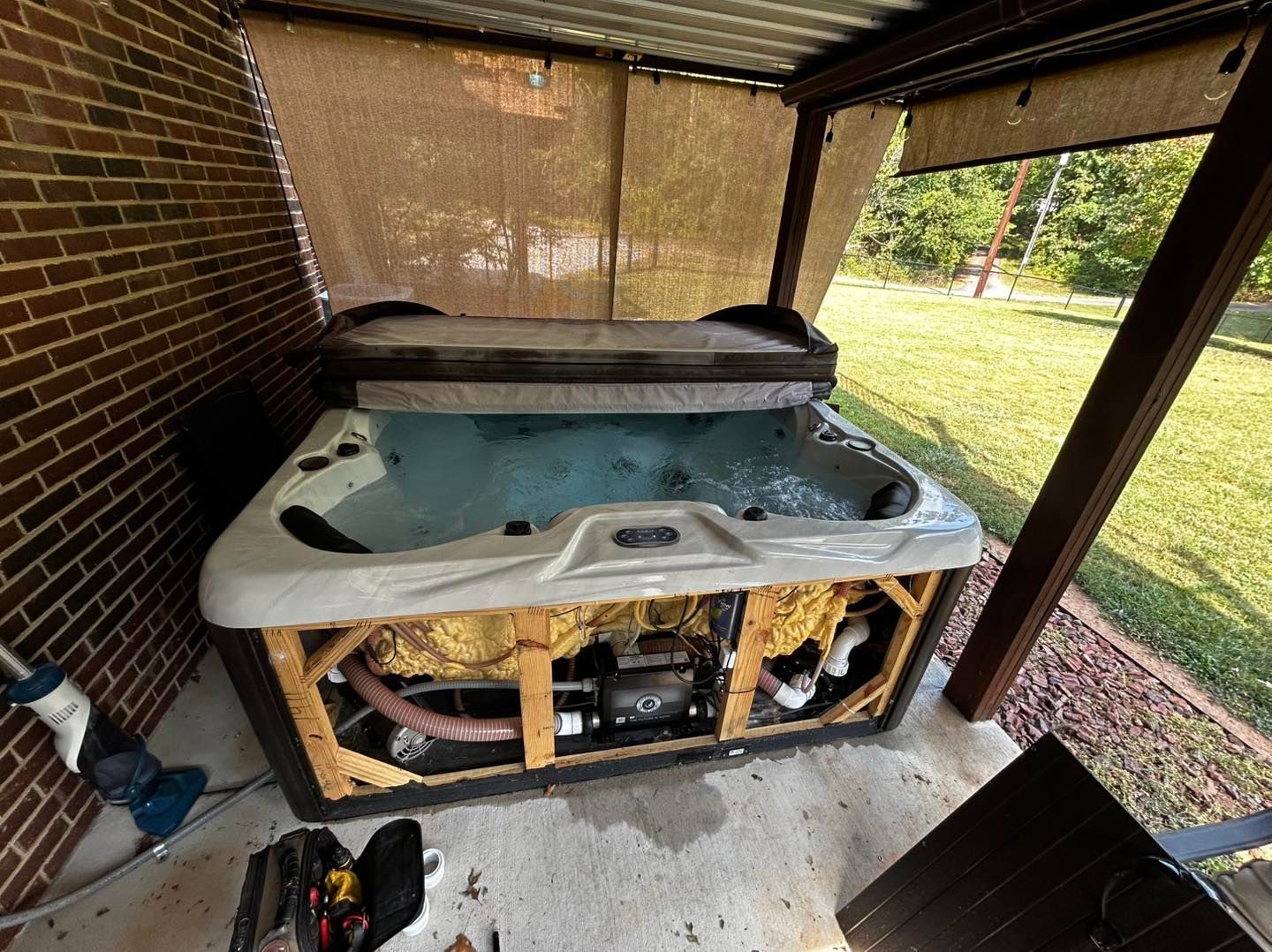 An outdoor hot tub with its access panel removed, revealing internal plumbing and pumps on a brick-walled patio.