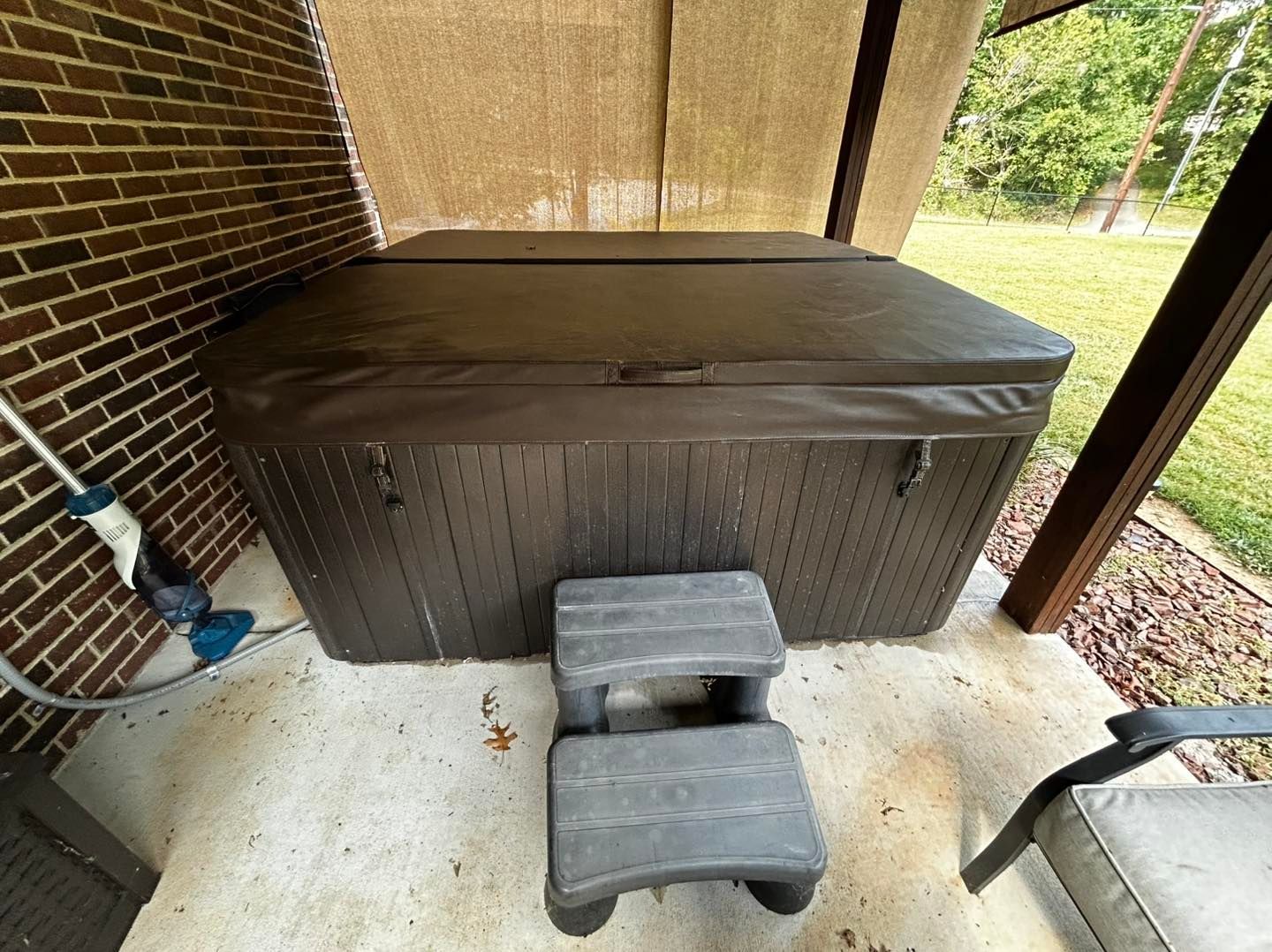 A dark brown hot tub with a cover sits on a concrete patio next to a brick wall, with a matching set of steps in front.