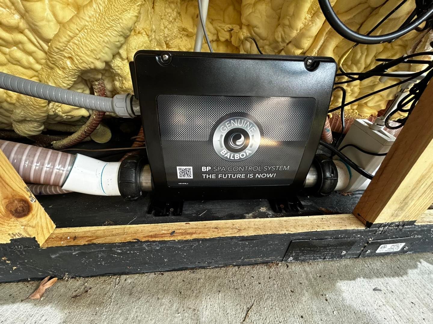 A black Balboa Water Group spa heater unit with connected piping, mounted in a wooden frame with yellow insulation.