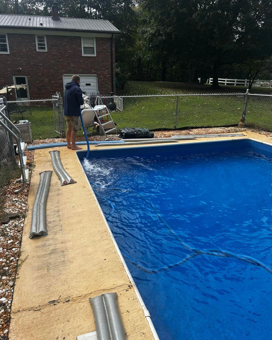 A person in a dark jacket uses a vacuum hose to clean a backyard swimming pool next to a brick house.