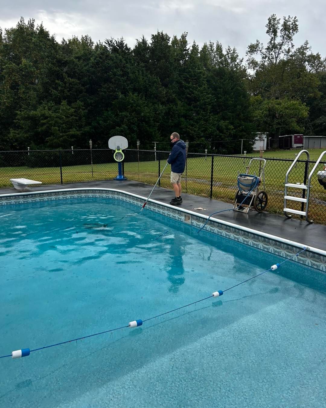 A person stands on a pool deck using a long-handled brush to clean the edge of a bright blue swimming pool.