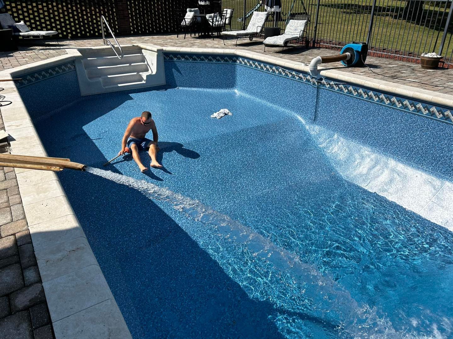 A person sits on the floor of an empty swimming pool while water flows from a hose nearby.