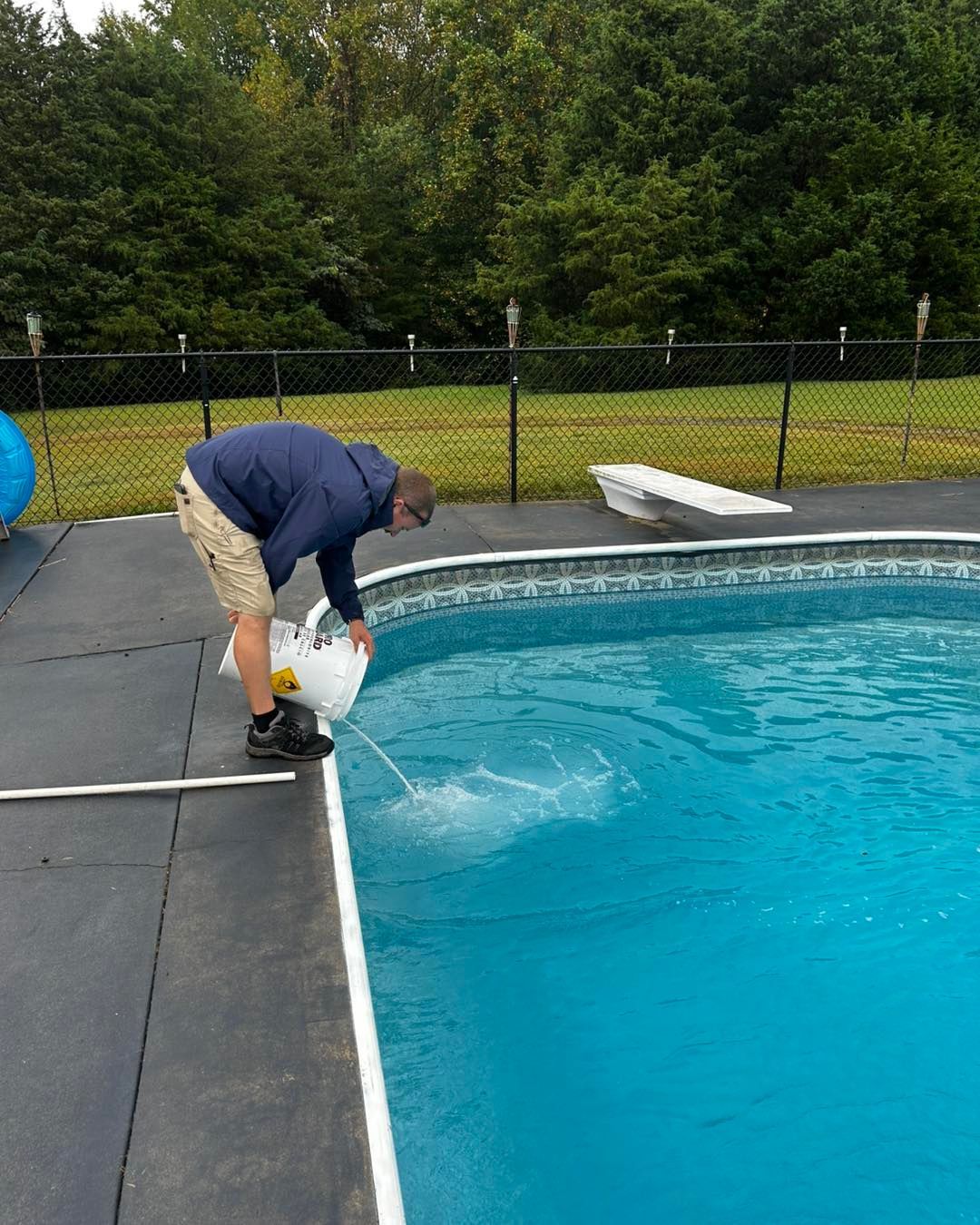 A person wearing a blue hoodie and khaki shorts pours a liquid from a white bucket into a backyard swimming pool.