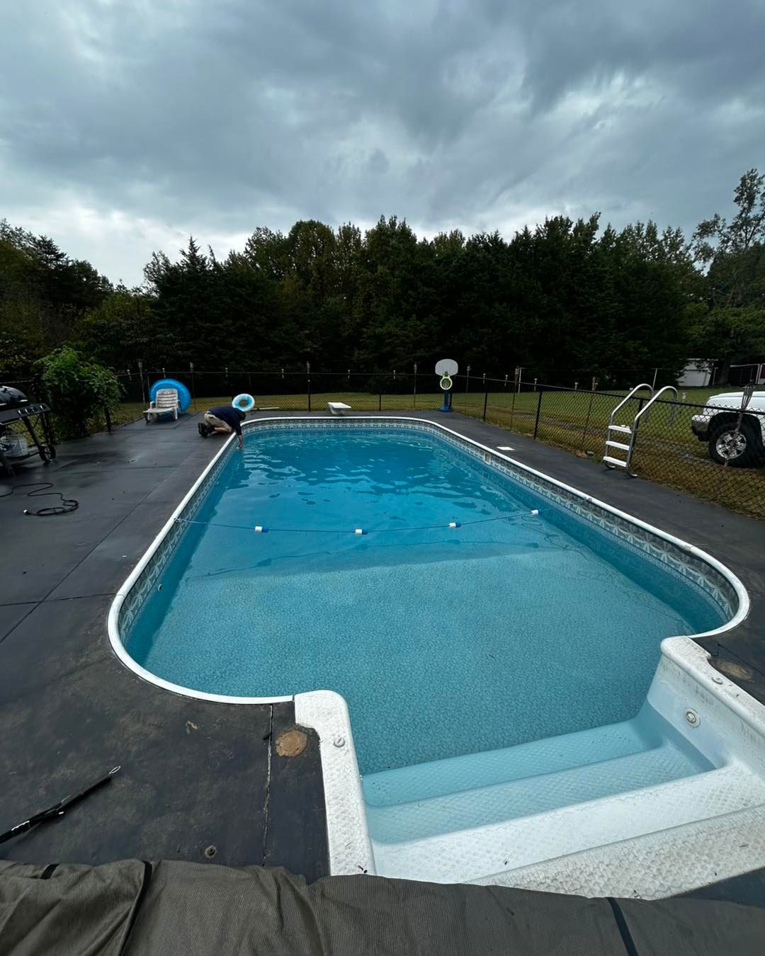 A rectangular swimming pool with a shallow end staircase, surrounded by a dark deck under a cloudy sky.