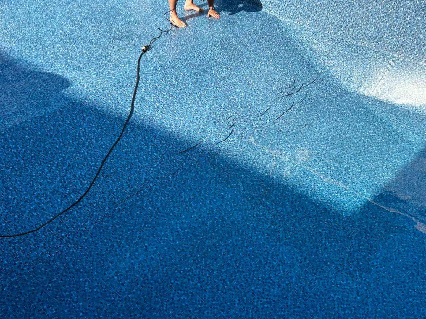 A person cleaning a blue speckled swimming pool floor with a vacuum hose, showing a clean path against dirty tile.