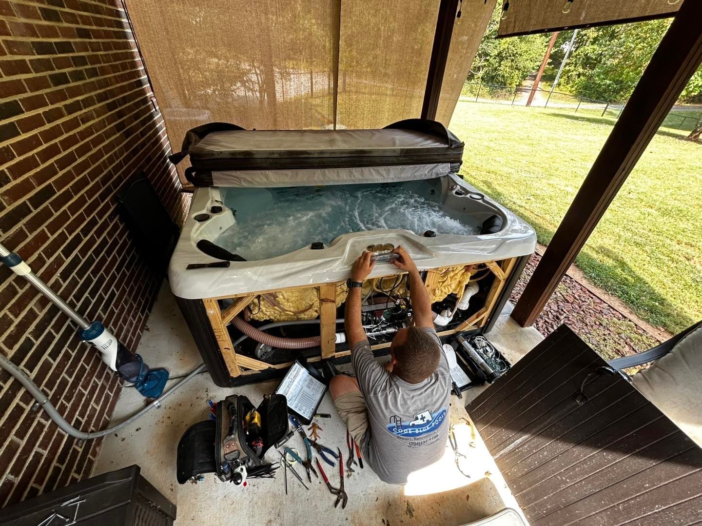 A technician repairs the exposed internal components of a hot tub on a brick-walled patio with tools spread nearby.