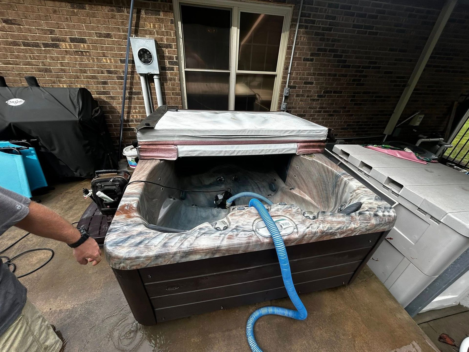 A blue hose drains a partially covered hot tub on a concrete patio next to a brick wall.