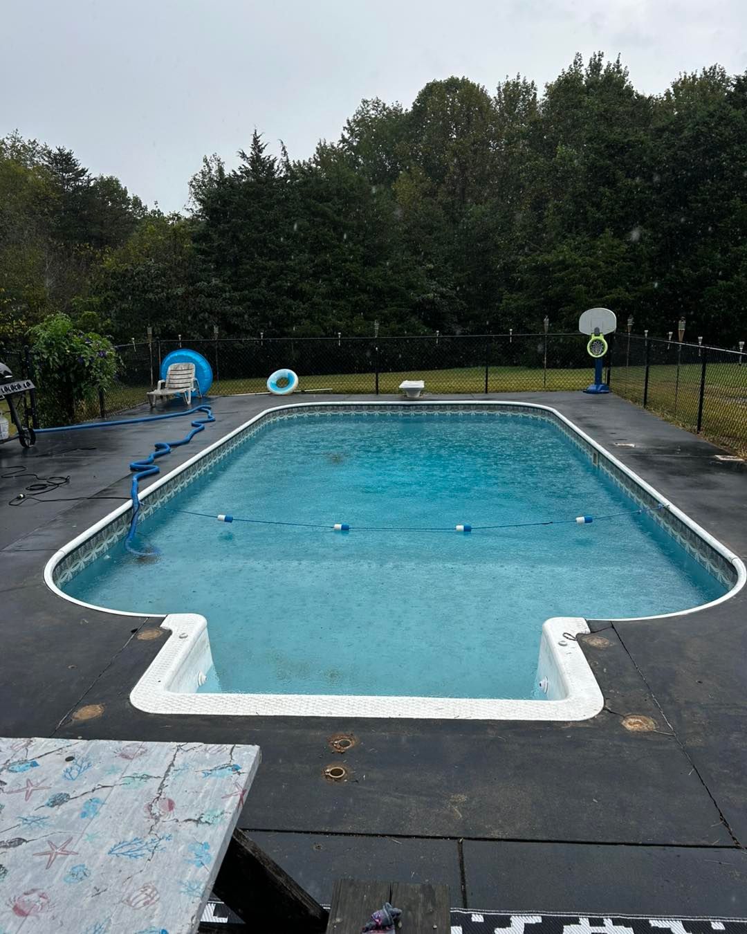 A rectangular swimming pool with an attached shallow area, surrounded by a dark concrete deck under a cloudy sky.