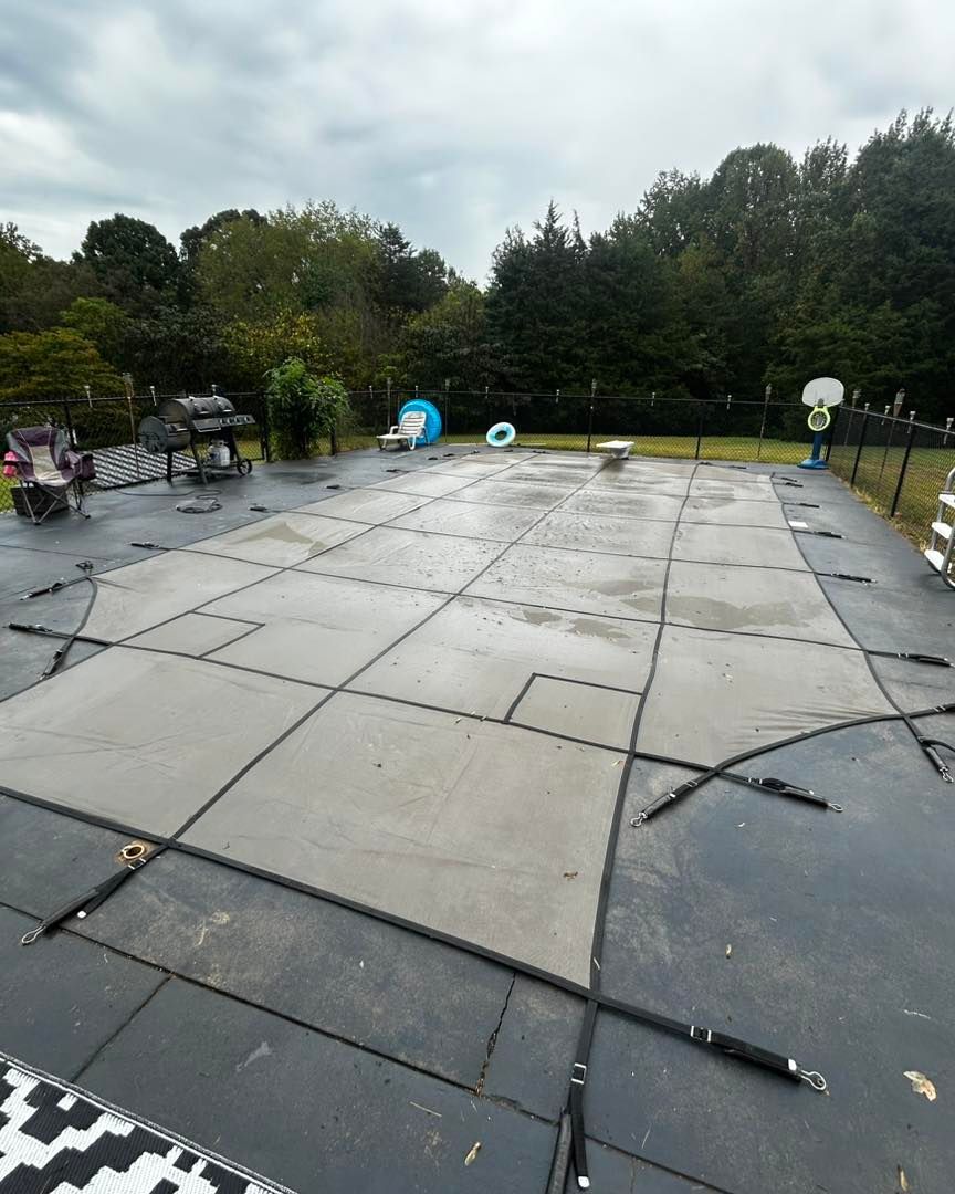 A rectangular swimming pool covered by a grey safety tarp, secured with straps on a dark patio in a backyard.
