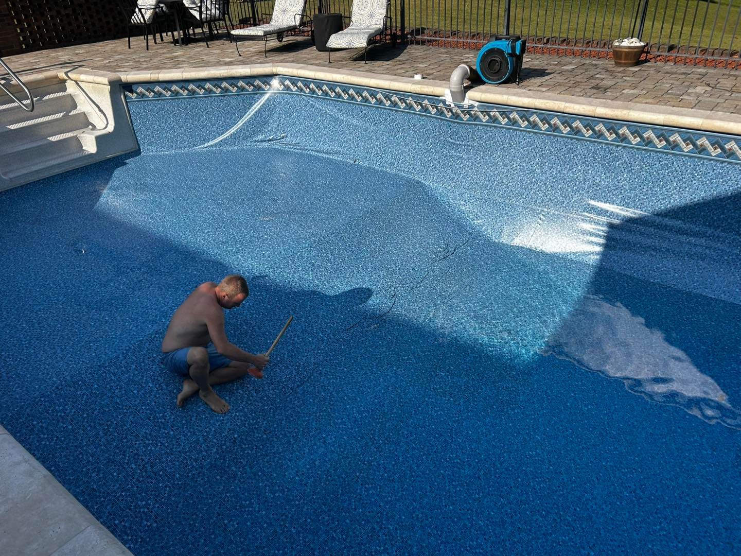 A person sits in the shallow end of a blue-tiled swimming pool, holding a long pole to clean the pool floor.