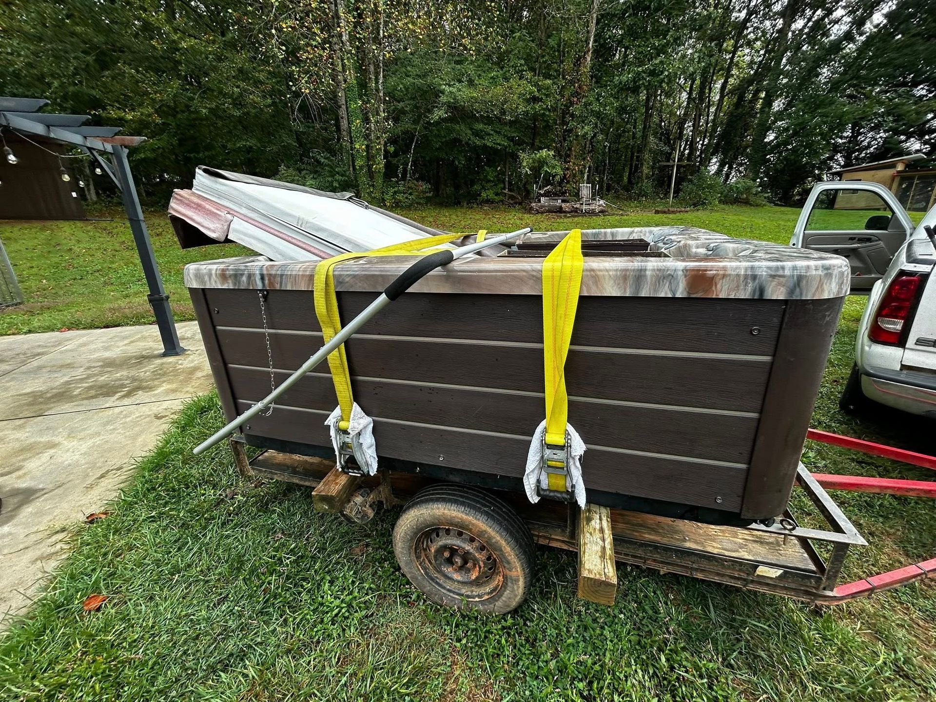 A brown hot tub is secured with yellow straps to a utility trailer parked on grass near a white vehicle.