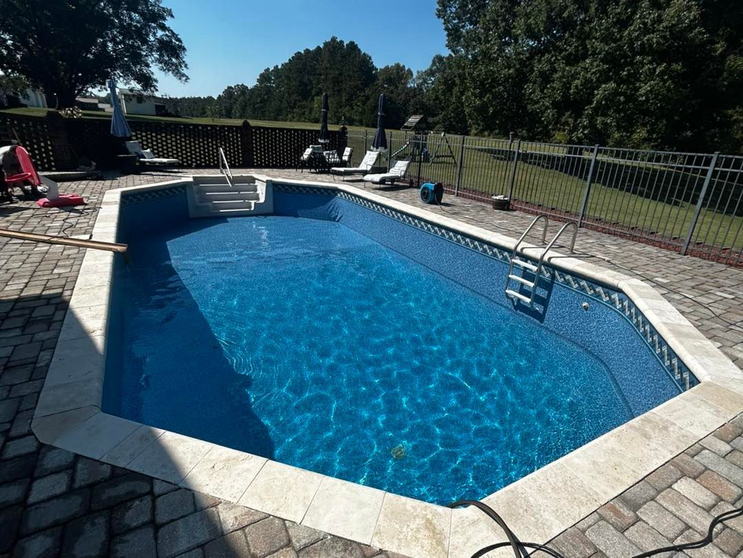 A backyard inground swimming pool with blue water and light stone coping, surrounded by a paved patio and black fence.