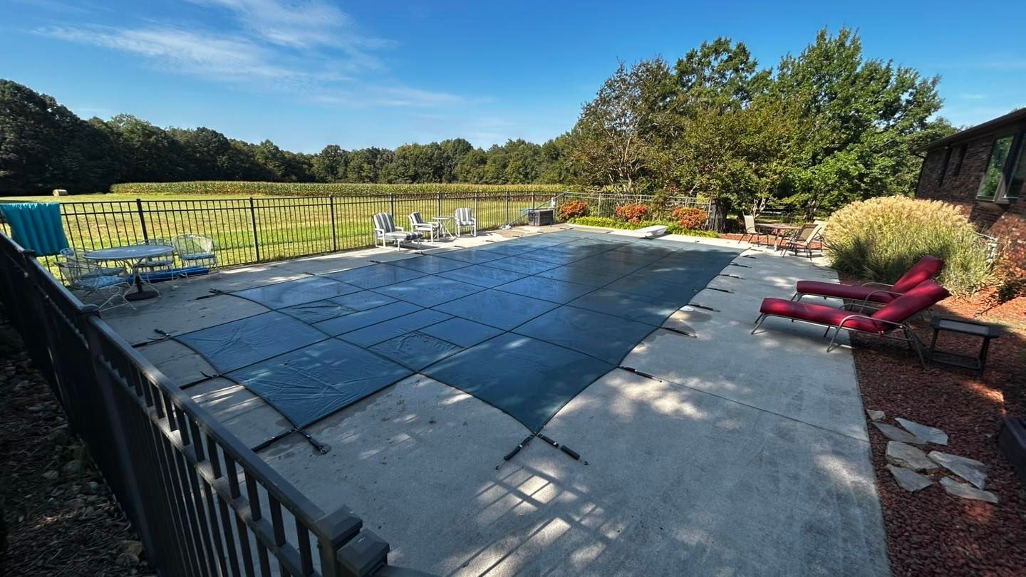 A rectangular swimming pool covered with a black safety net, surrounded by concrete decking and red lounge chairs.
