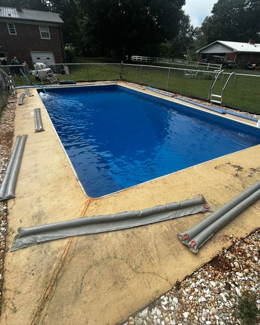 A bright blue rectangular swimming pool surrounded by a tan concrete deck with gray water bags laid out for a pool cover.