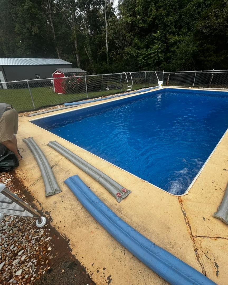 A rectangular swimming pool with deep blue water, surrounded by a light-colored patio with scattered pool cover weights.
