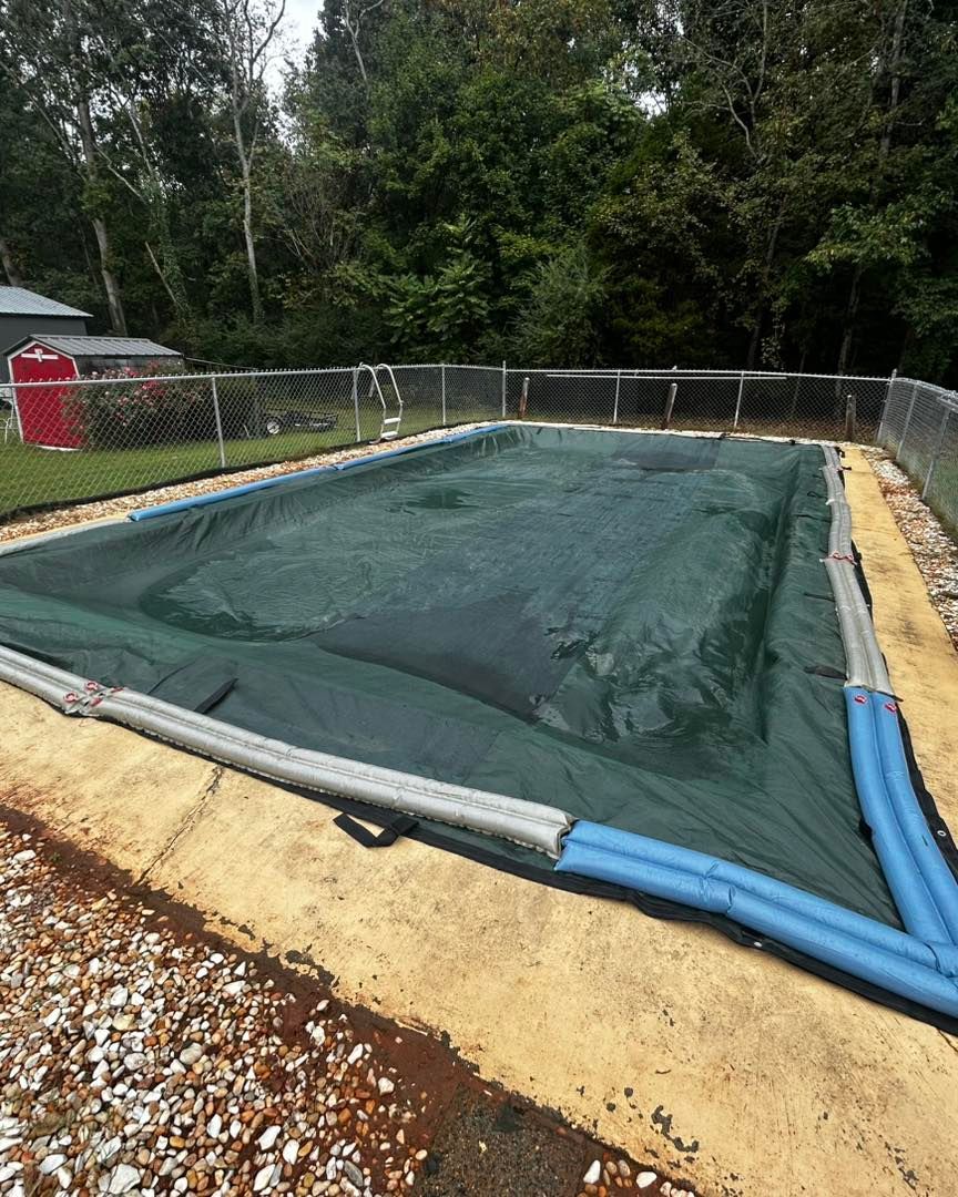 A rectangular in-ground pool covered with a dark green tarp and secured by blue water tubes on a concrete deck.