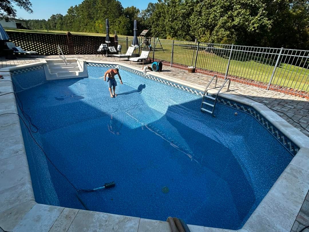 A person stands inside an empty, blue, tiled swimming pool on a sunny day with a fence and trees in the background.