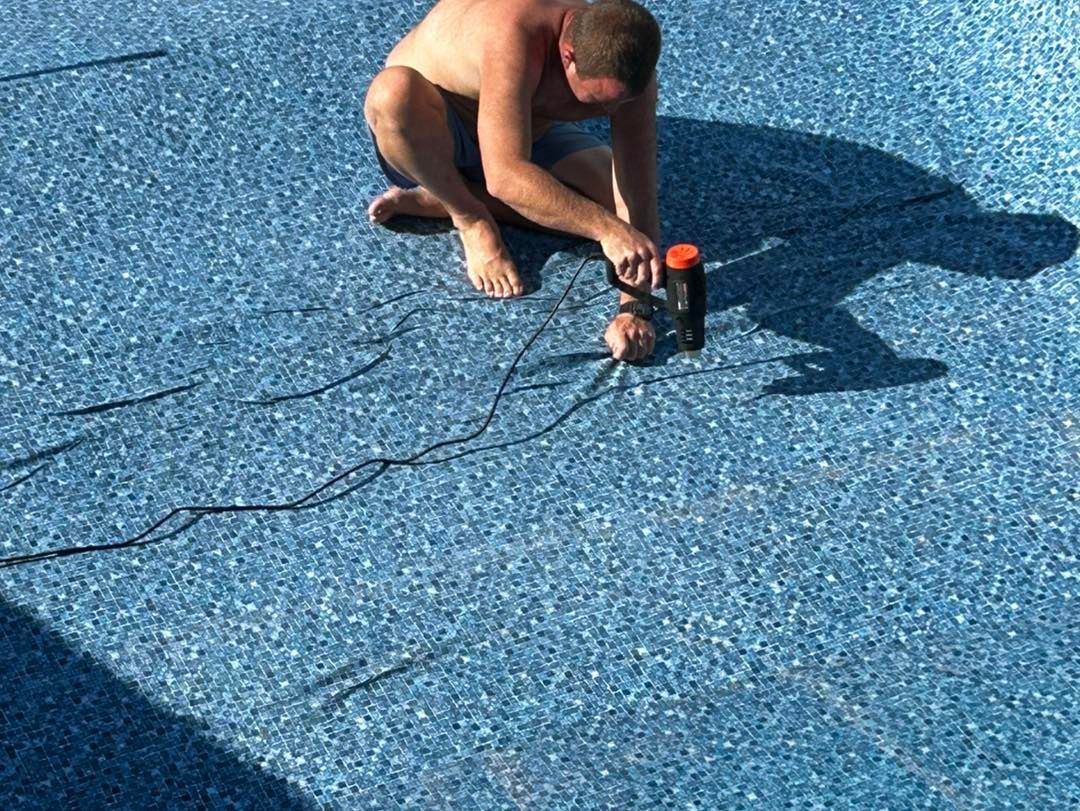 A person kneeling in an empty swimming pool uses a heat gun to seal a seam on the blue patterned vinyl liner.