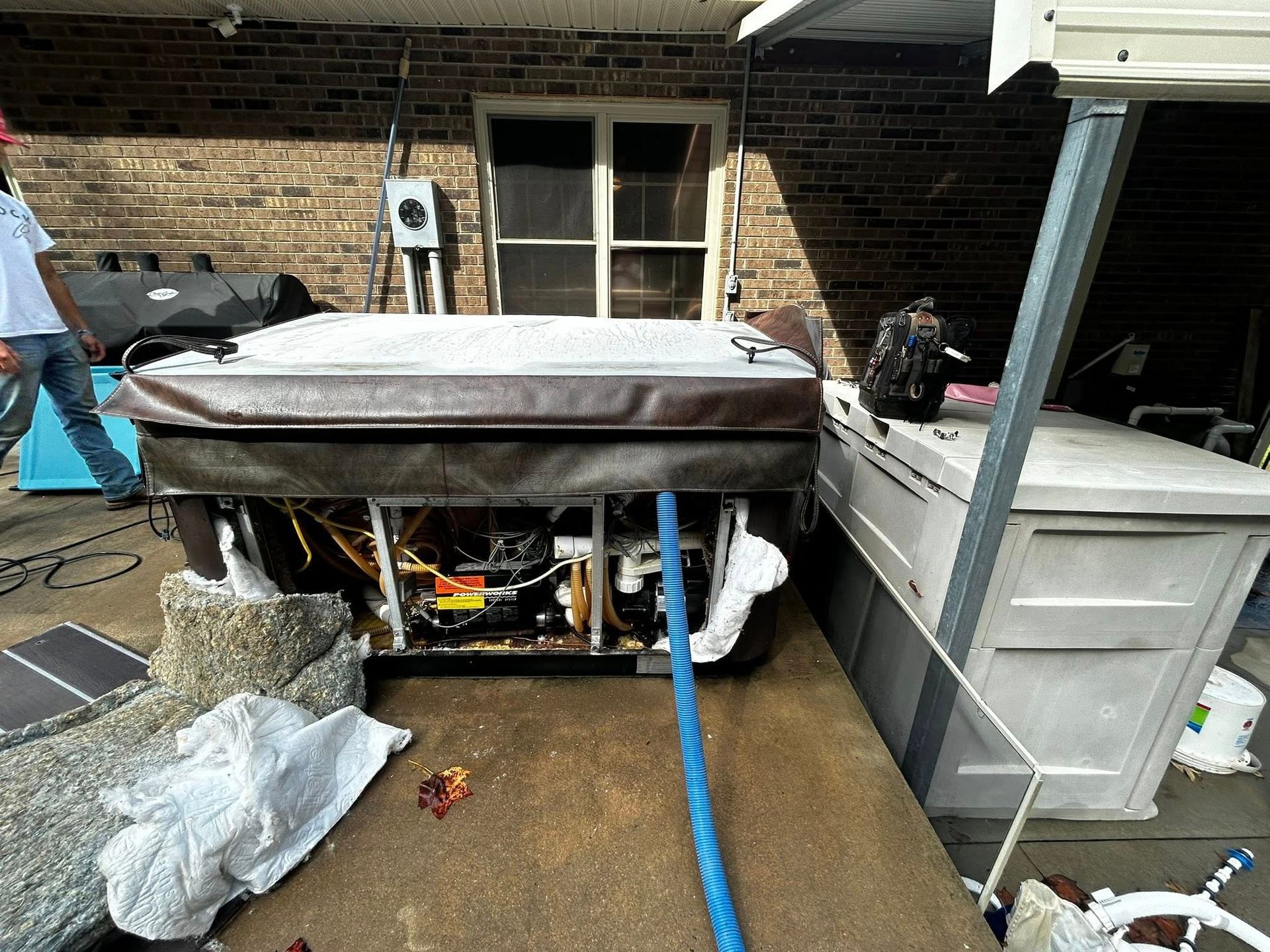 A hot tub with an open side panel sits on a concrete patio next to a person and a white plastic storage shed.