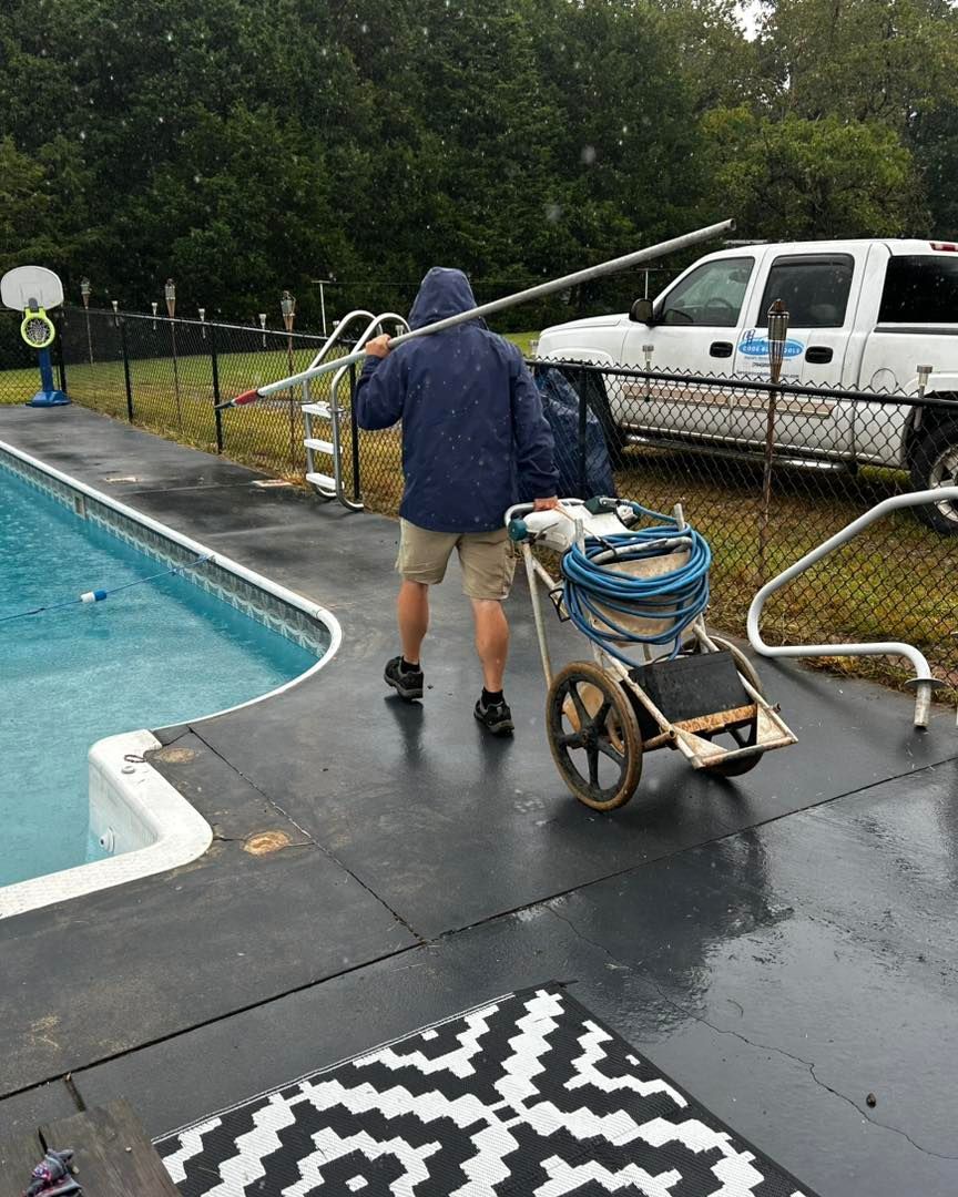 A service professional carries a long pole and wheels equipment along a wet pool deck near a service truck.