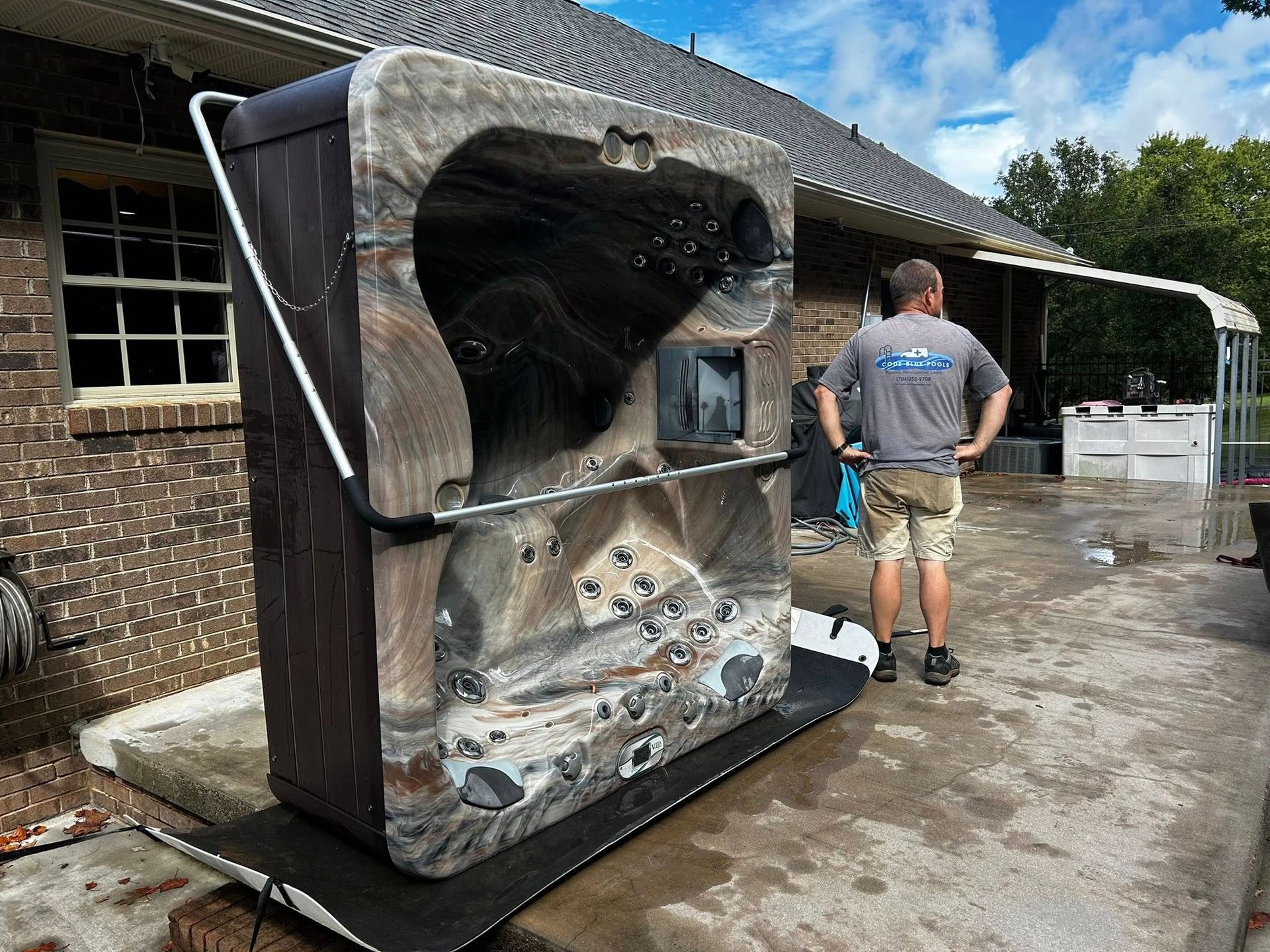 A person in a gray shirt stands next to a hot tub turned on its side on a black platform outside a brick house.