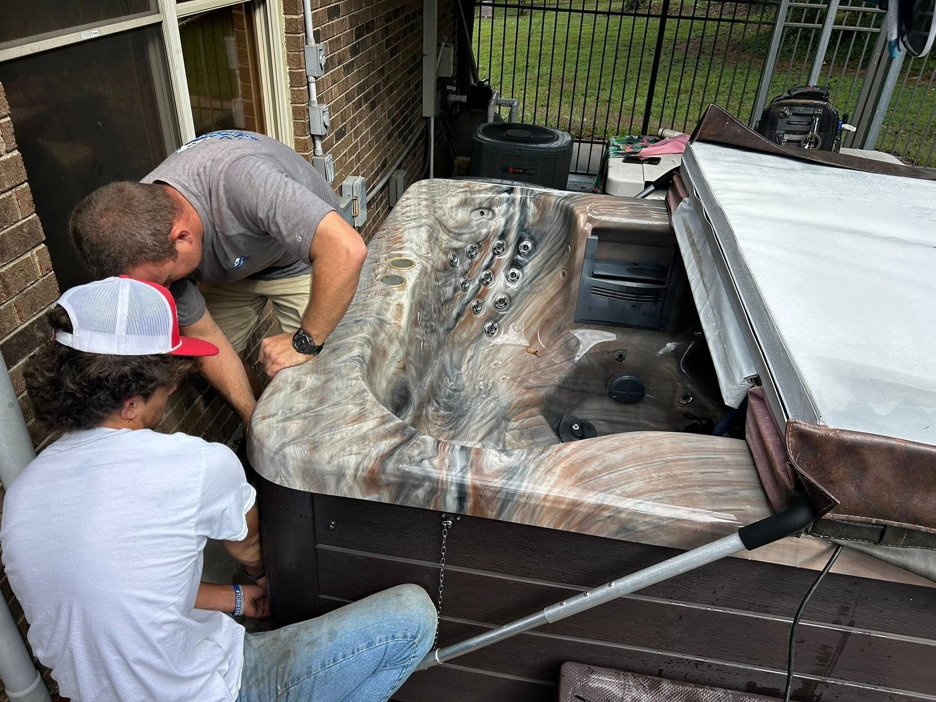 Two people work on a hot tub with a marbled interior on an outdoor patio near a brick wall.