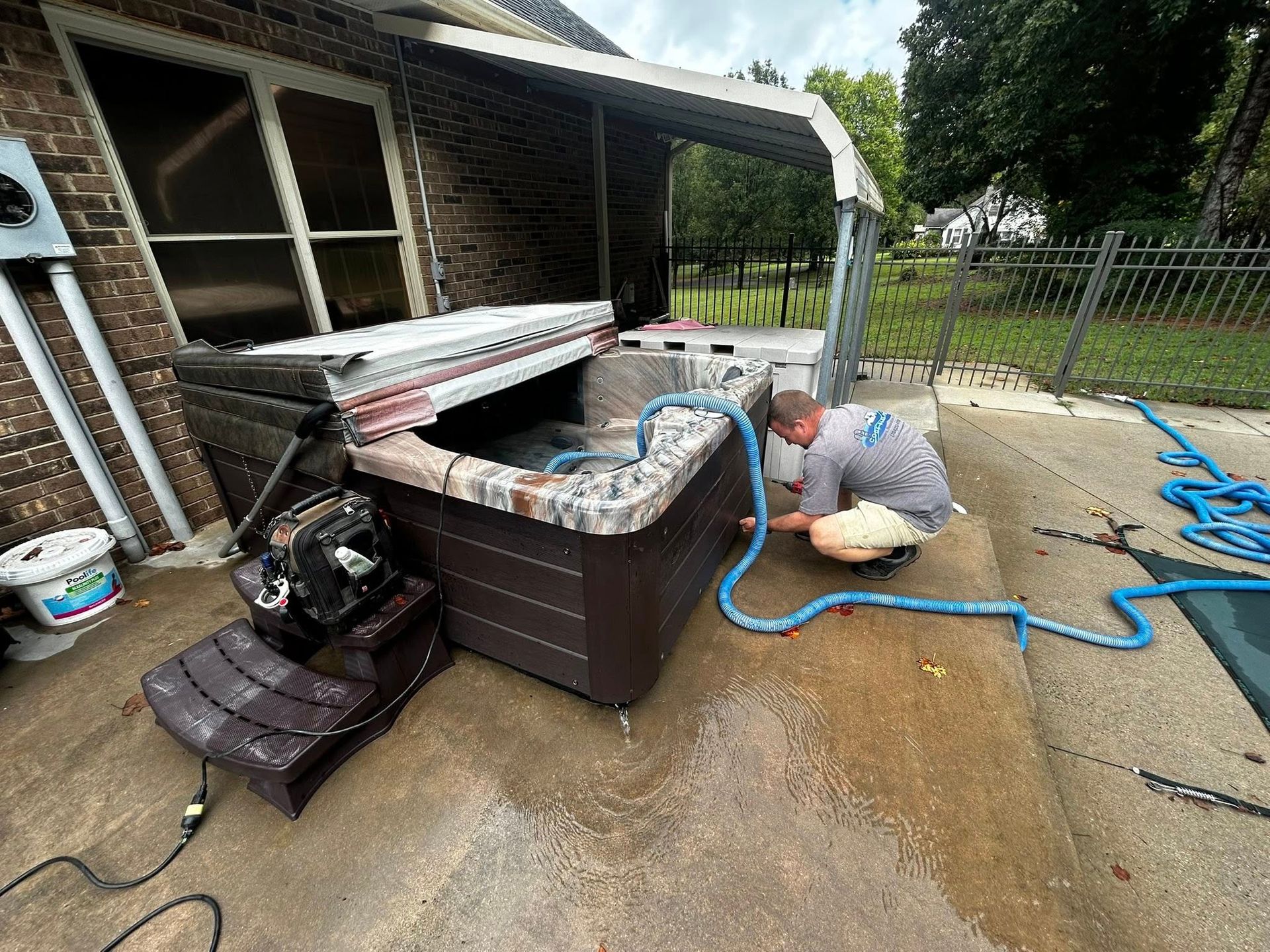 A person in a gray shirt kneeling to repair a brown hot tub outdoors on a concrete patio.