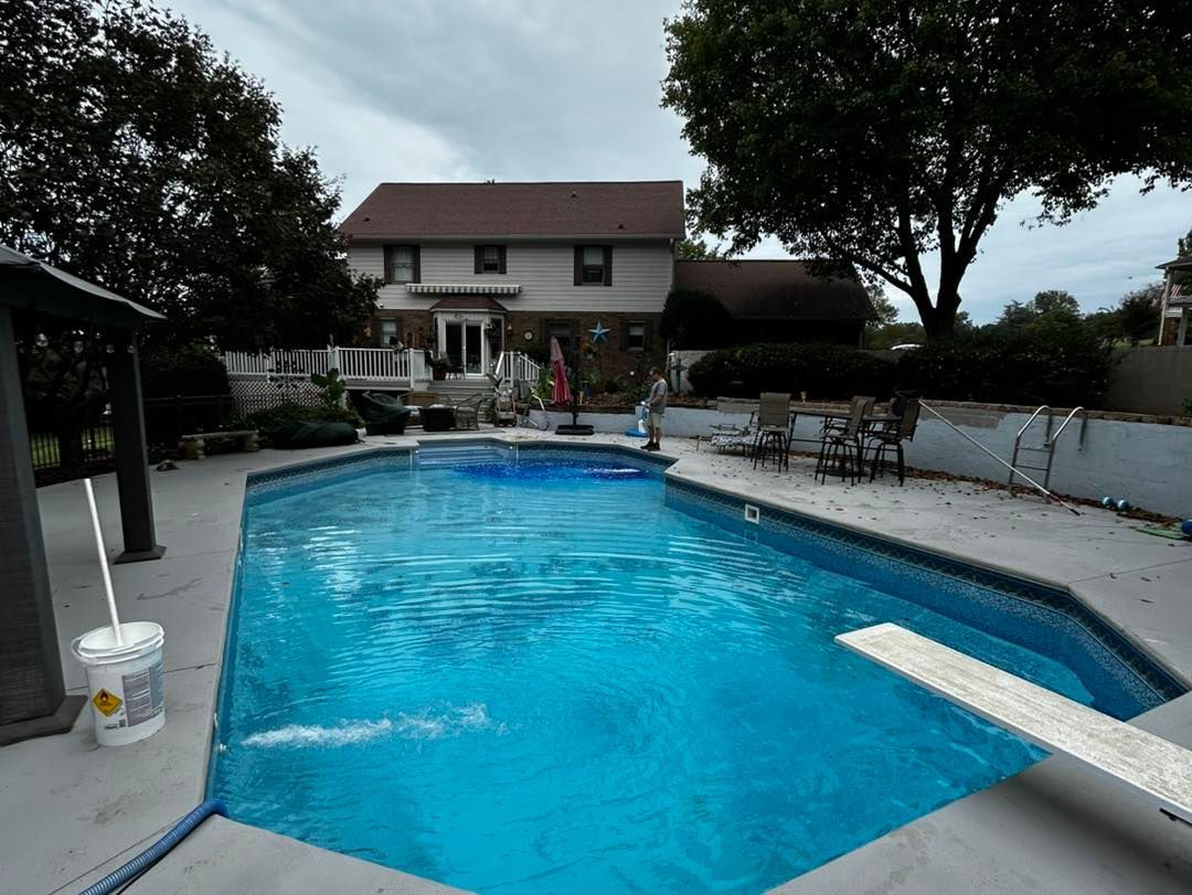 A backyard swimming pool with a diving board and patio, located in front of a two-story suburban house.
