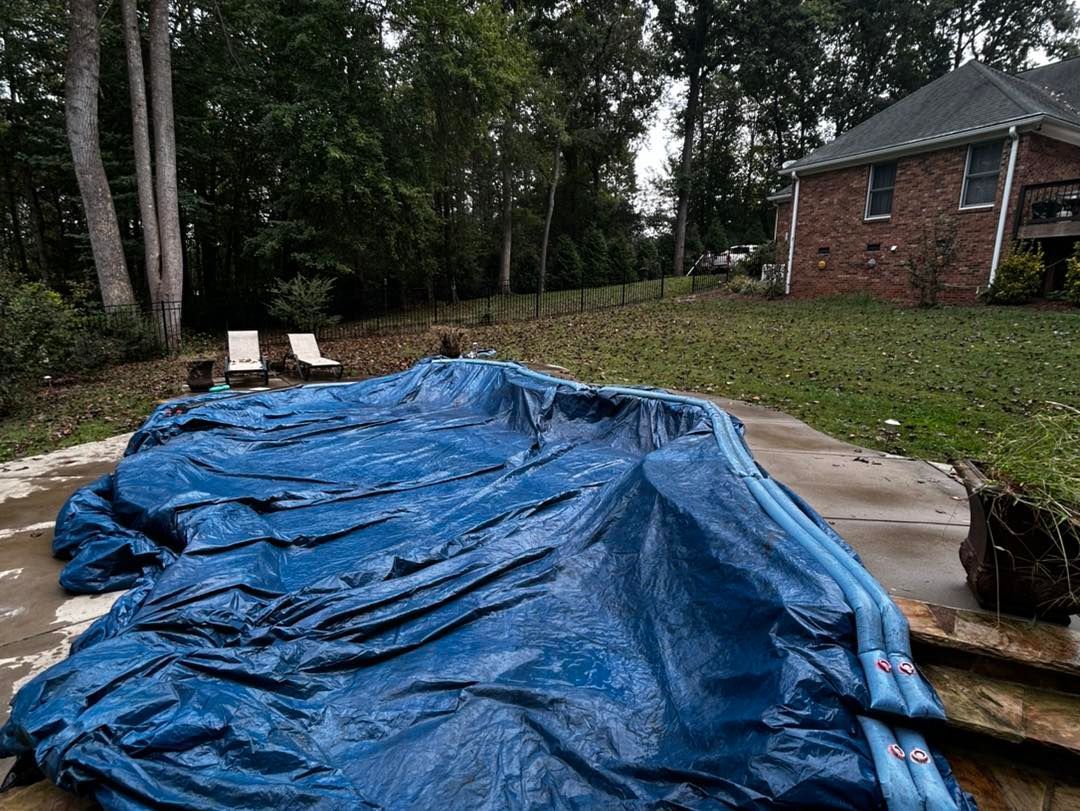 A blue pool cover lies over an in-ground pool in a backyard near a brick house and two lounge chairs.
