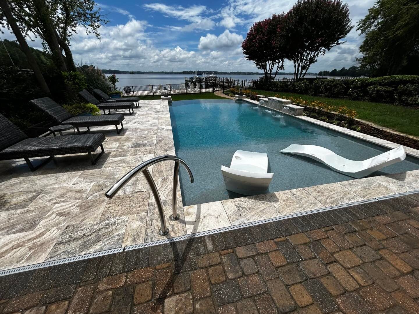 A swimming pool with two lounge chairs in shallow water, surrounded by a stone deck overlooking a lake on a sunny day.