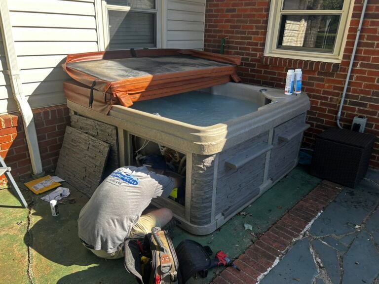 A technician kneels to repair the exposed equipment compartment of an outdoor hot tub on a brick patio.