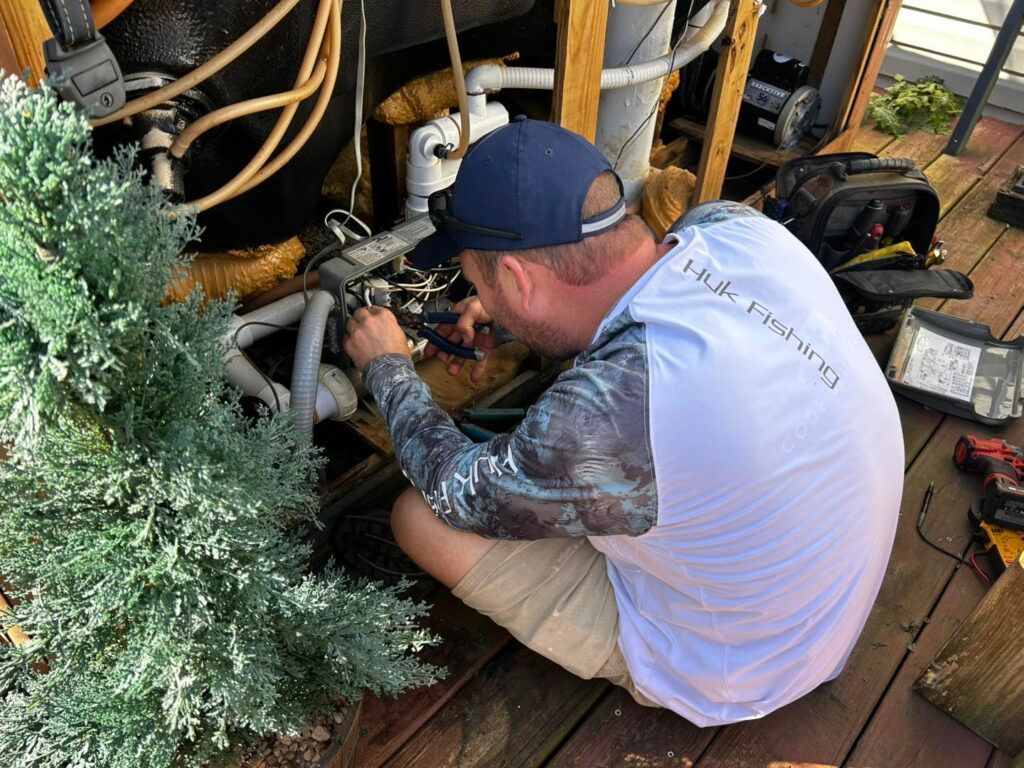A person in a white and patterned long-sleeve shirt kneels on a deck while repairing plumbing under a hot tub.