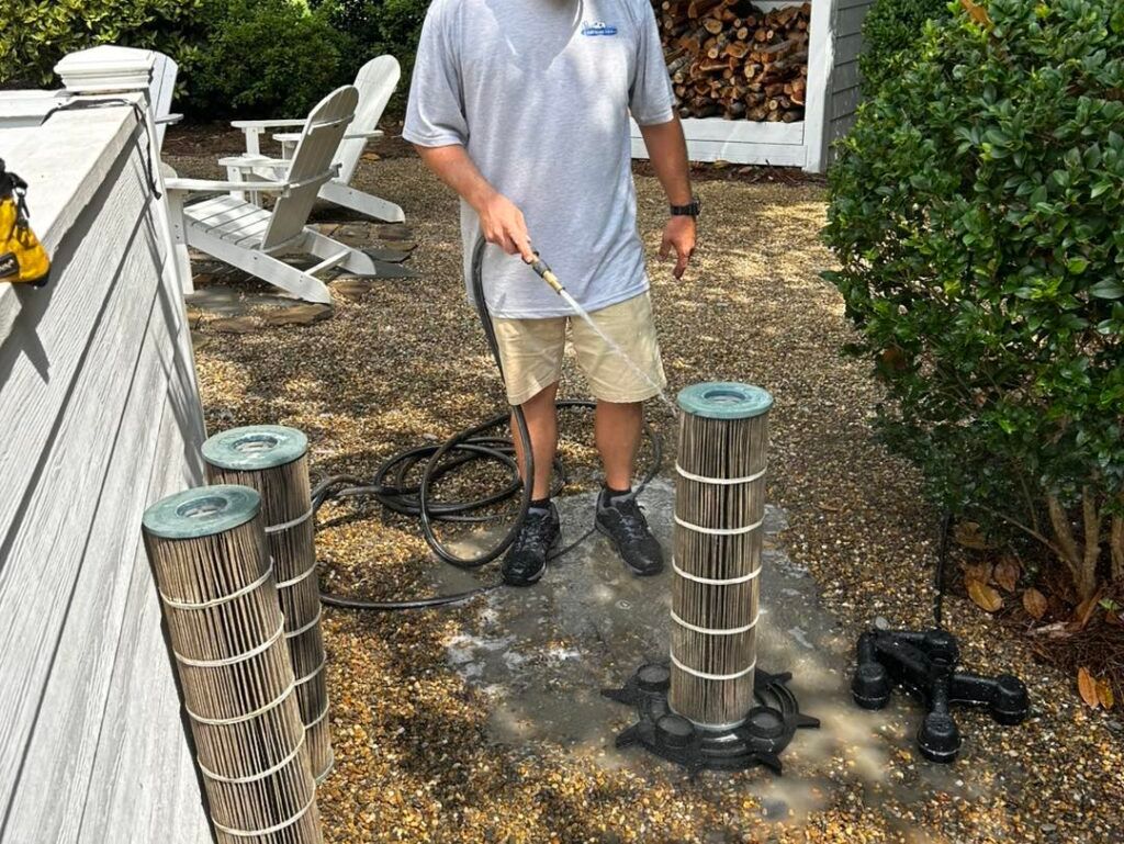 A person sprays water from a hose onto vertical pool filter cartridges standing on a gravel patio.