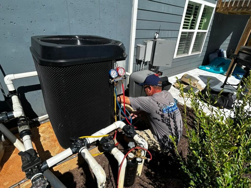 A technician in a gray shirt services an outdoor pool heater, using manifold gauges connected to the system.