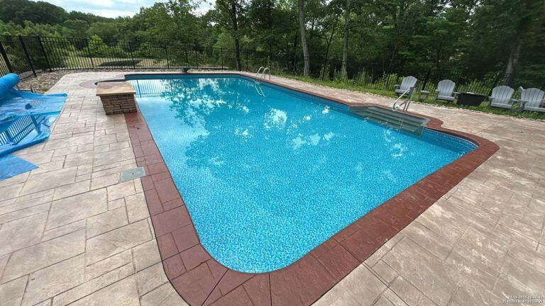 A rectangular outdoor swimming pool with a reddish-brown border surrounded by light stone pavers and trees.