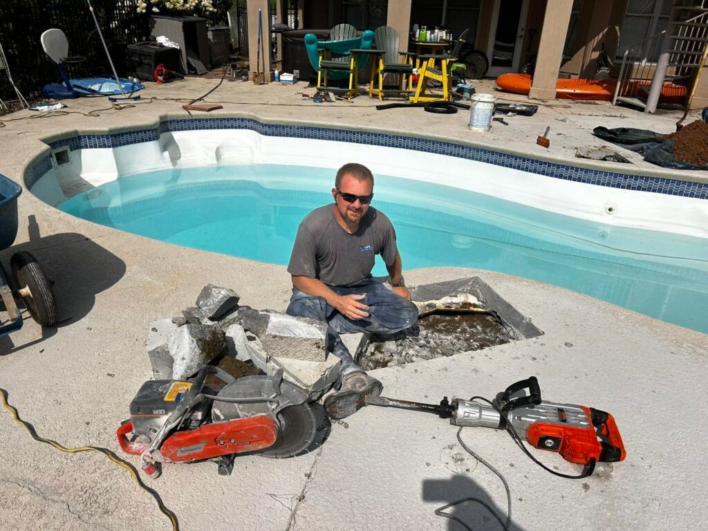 A person in a gray shirt kneeling by a pool deck next to a concrete saw and power tool after cutting a hole in the ground.