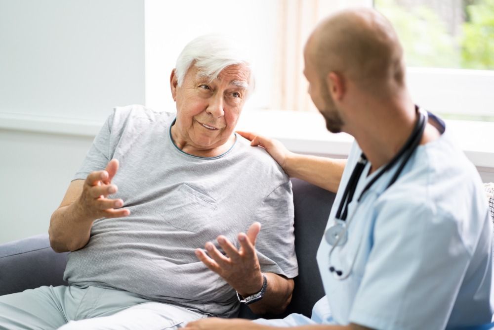 An elderly man is sitting on a couch talking to a doctor.