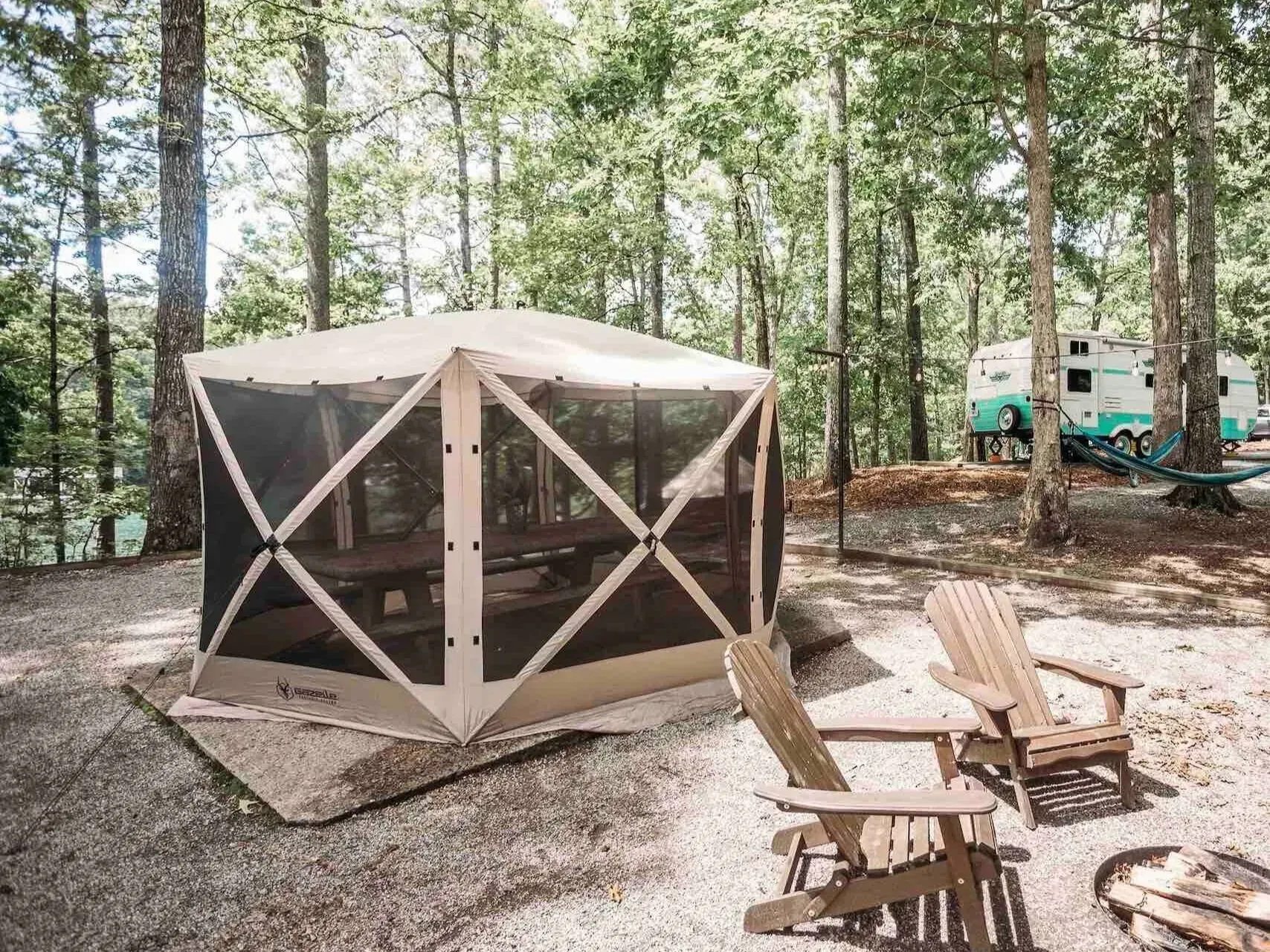Screened camping shelter with chairs on gravel, trees in background, and a vintage camper.