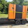 An orange and black Elgi air compressor machine on a flatbed trailer outdoors. The machine is secured with yellow strapping.