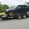 Dark blue SUV on a black trailer, parked on a gray road near a green lawn and trees.