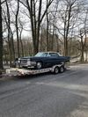 A classic dark blue car on a trailer, parked on a gravel road, surrounded by trees.