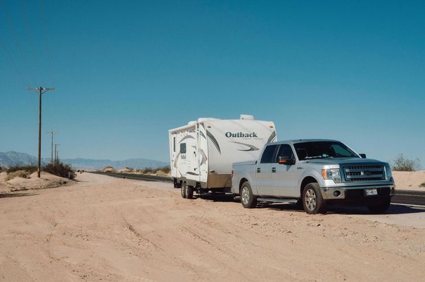 Silver pickup truck towing a white RV on a dirt road under a clear blue sky. Desert landscape surrounds them.